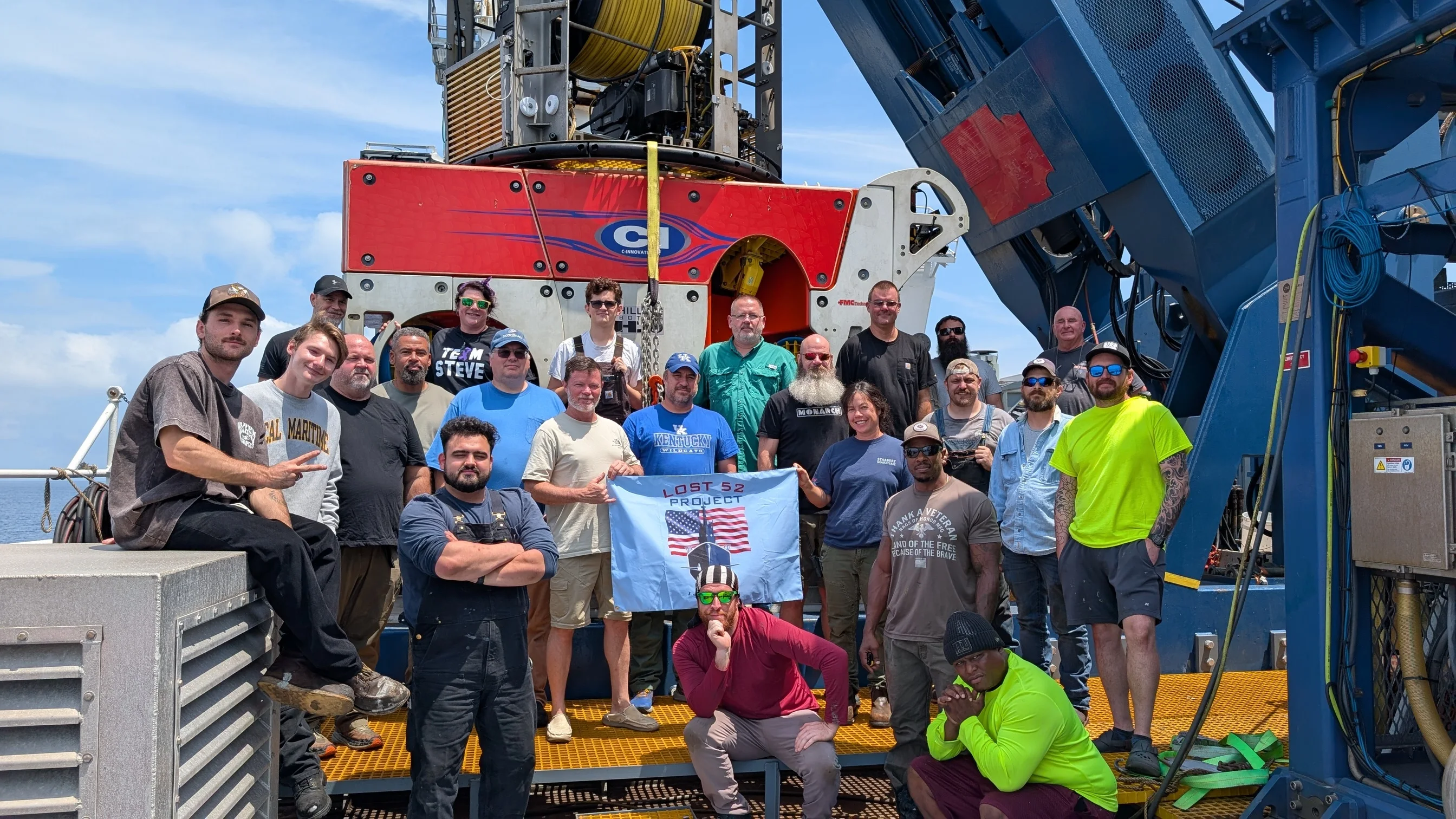 The 2025 and 2026 discovery team on deck with the ROV, holding a Lost 52 Project flag
